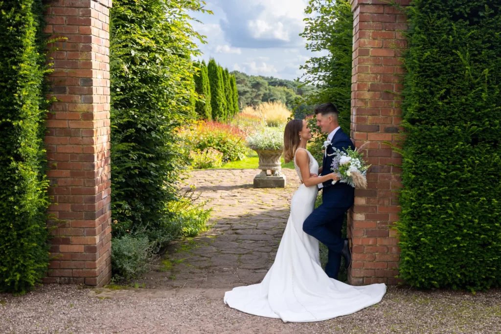A bride and groom stand facing each other in a garden, surrounded by brick pillars and greenery, with the bride holding a bouquet of flowers. Copyright Somerside Photography Ltd // www.somersidephotography.com