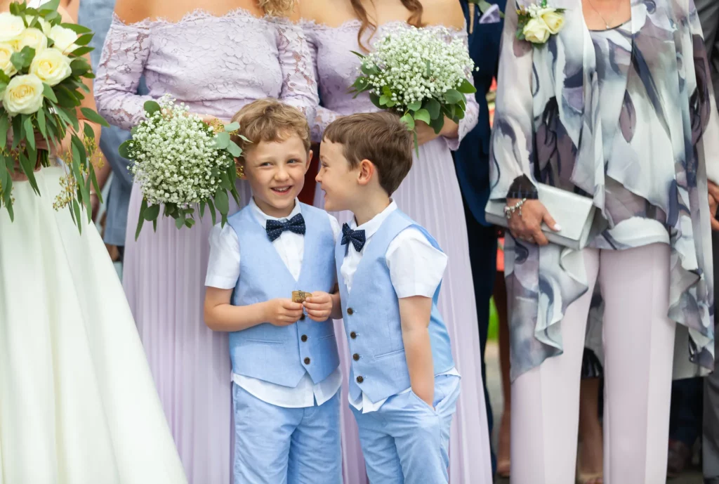 Two young boys in matching light blue suits stand together at a wedding, surrounded by women holding bouquets and wearing lavender and floral dresses. Copyright Somerside Photography Ltd // www.somersidephotography.com