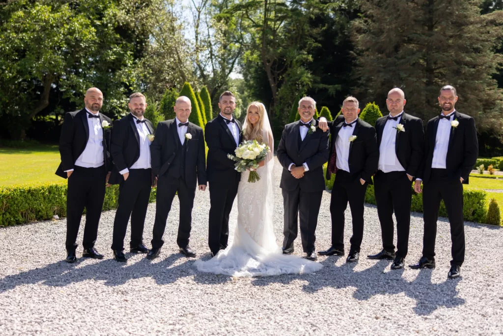A bride and groom stand outdoors in the centre of a group of men in tuxedos, all posing and smiling for a formal wedding photo on a gravel path with greenery in the background. Copyright Somerside Photography Ltd // www.somersidephotography.com