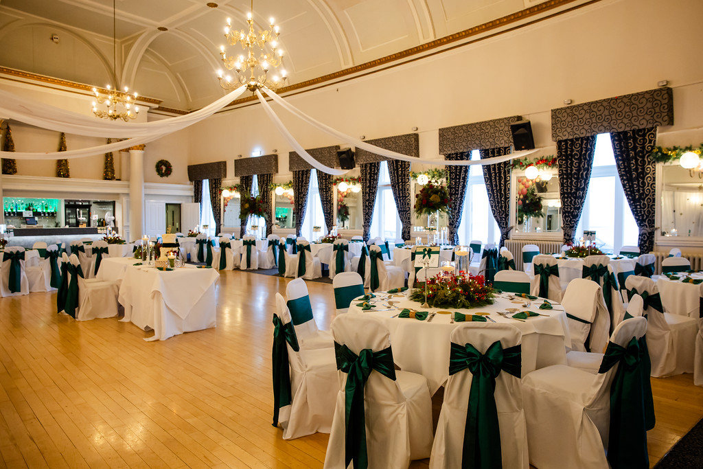 A formal event hall with round tables covered in white cloths and dark green chair sashes, set for a banquet, with chandeliers and draped white fabric on the ceiling. Copyright Somerside Photography Ltd // www.somersidephotography.com