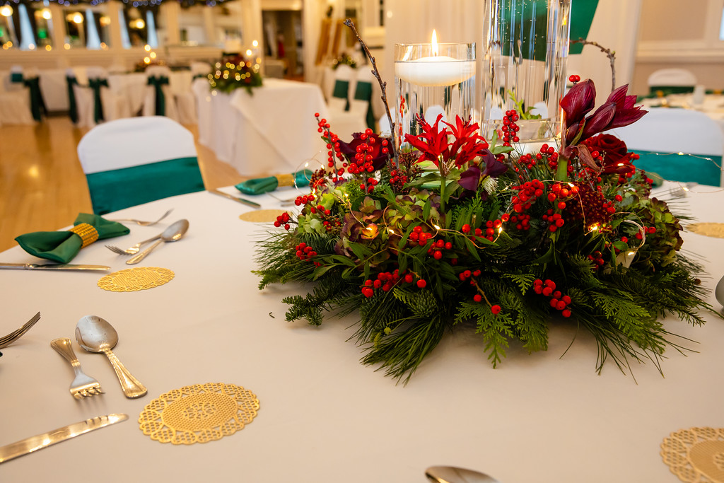 A round table set for a formal event features a festive centrepiece with red flowers, berries, greenery, and a tall glass candle. Gold coasters and cutlery are arranged neatly around the table. Copyright Somerside Photography Ltd // www.somersidephotography.com