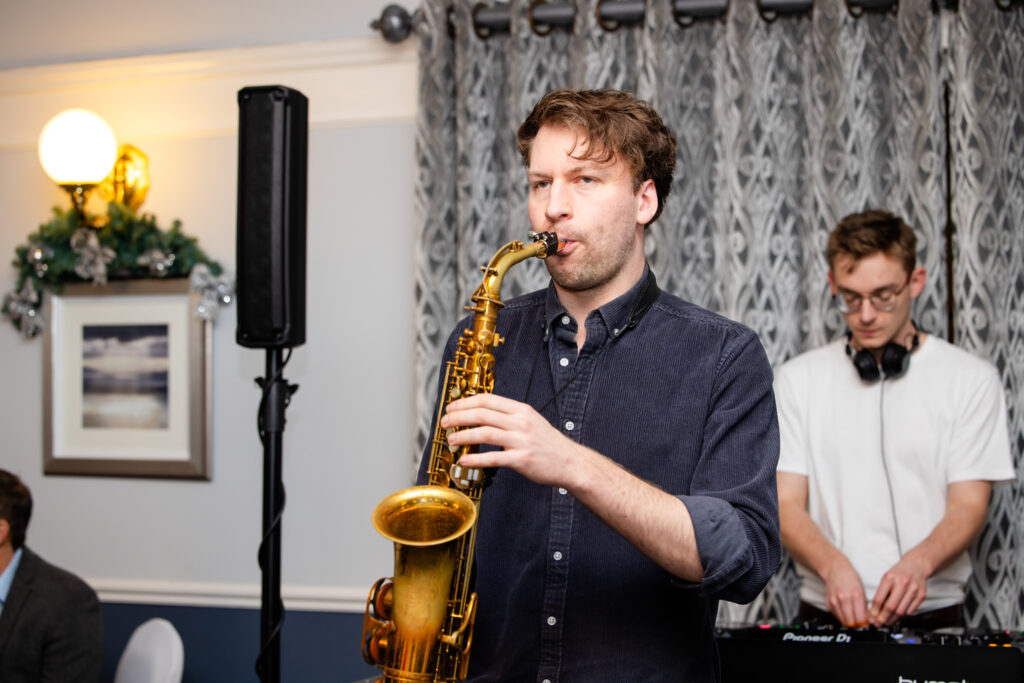 A man plays a saxophone whilst another man operates DJ equipment in a room decorated with patterned curtains and a wall light. Copyright Somerside Photography Ltd // www.somersidephotography.com