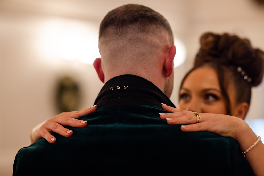A woman with manicured nails and a ring embraces a man in a dark velvet jacket with “14.12.24” embroidered on the collar. Copyright Somerside Photography Ltd // www.somersidephotography.com