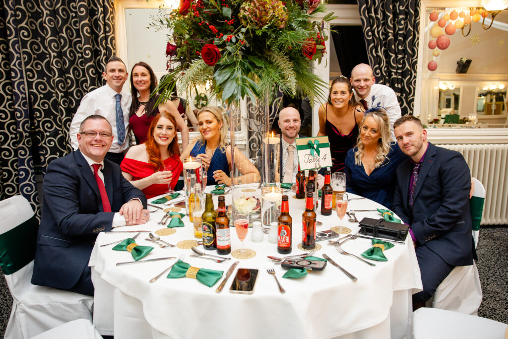 A group of people dressed formally are seated around a decorated banquet table with drinks and flowers at an indoor event. Copyright Somerside Photography Ltd // www.somersidephotography.com