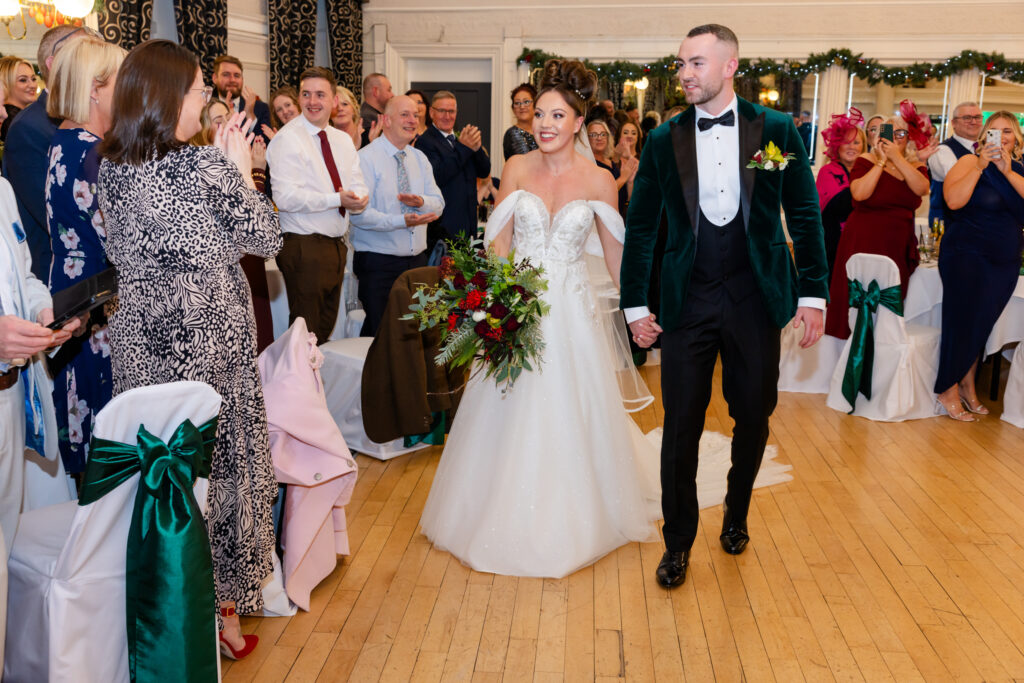 A bride and groom walk hand-in-hand down the aisle at their wedding, surrounded by guests standing and clapping in a decorated indoor venue. Copyright Somerside Photography Ltd // www.somersidephotography.com