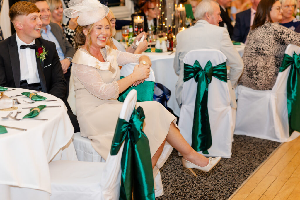 Guests at a formal event sit at tables with white cloths and green chair sashes. A smiling woman in a cream dress and hat sits in the foreground. Copyright Somerside Photography Ltd // www.somersidephotography.com