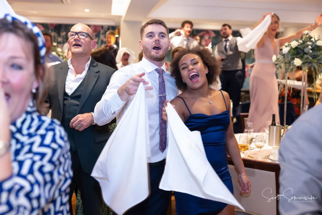 Guests at a formal event enthusiastically wave serviettes and sing, with tables and floral decorations visible in the background. Copyright Somerside Photography Ltd // www.somersidephotography.com