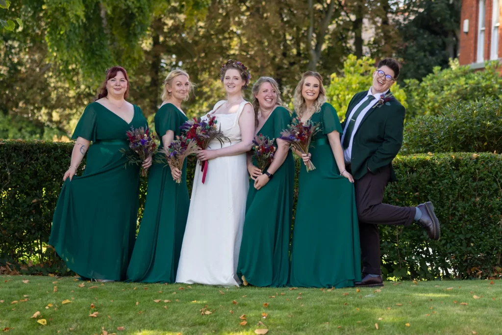 Six people posing outdoors; one in a white wedding dress and five in matching green dresses or suits, all holding bouquets, smiling, and standing on grass with trees and hedges in the background. Copyright Somerside Photography Ltd // www.somersidephotography.com