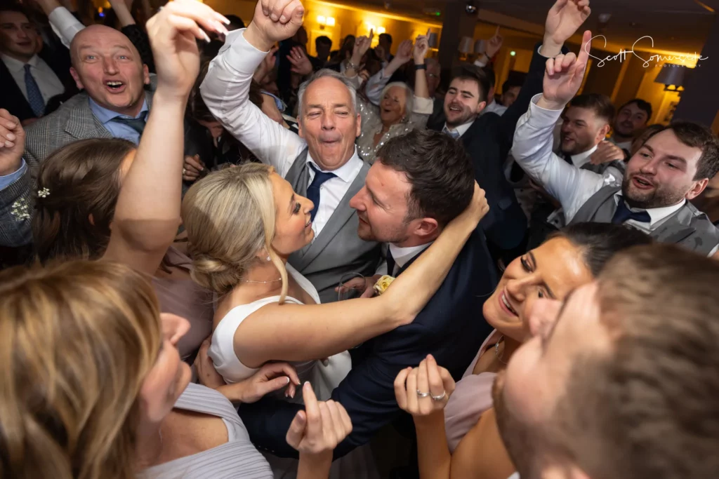 A bride and groom embrace and smile surrounded by joyful guests dancing and celebrating at a wedding reception. Copyright Somerside Photography Ltd // www.somersidephotography.com