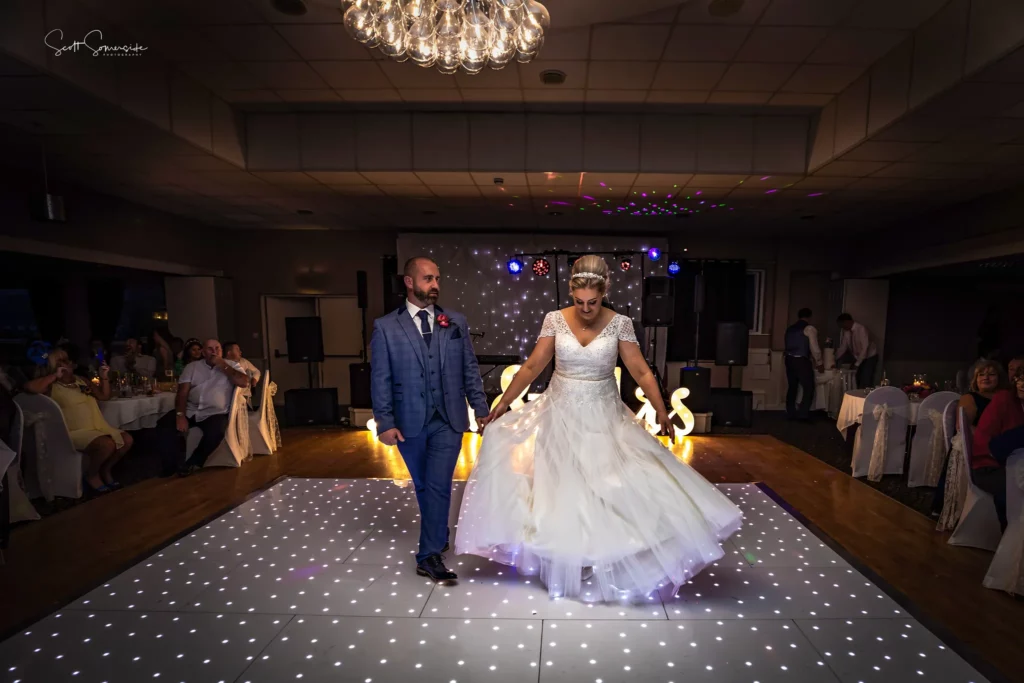 A bride and groom dance on a lit-up floor in a decorated reception hall, surrounded by seated guests, under a chandelier. Copyright Somerside Photography Ltd // www.somersidephotography.com