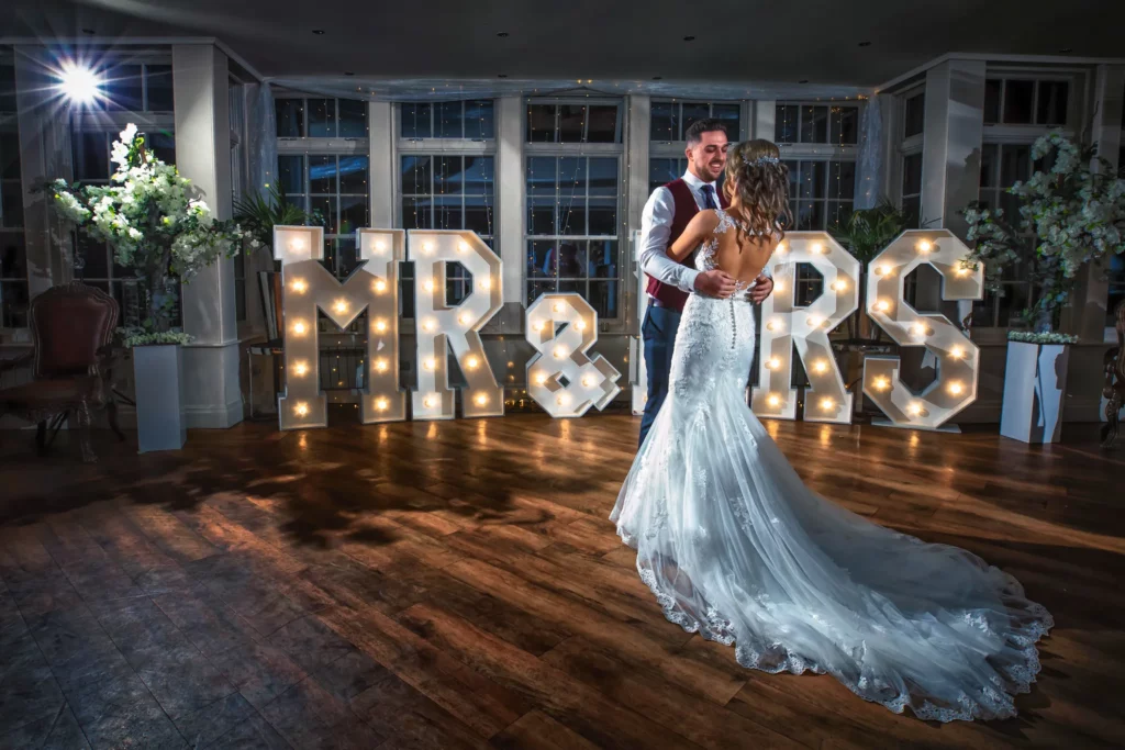 A bride and groom share their first dance in front of illuminated "MR & MRS" letters in a decorated reception hall. Copyright Somerside Photography Ltd // www.somersidephotography.com