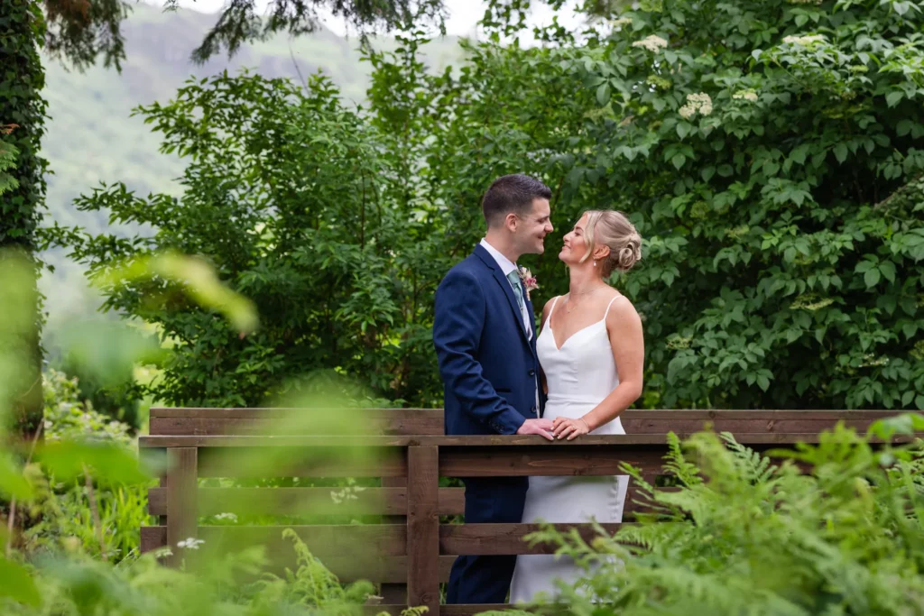 A couple dressed in formal wedding attire stand together on a wooden bridge surrounded by lush green foliage. Copyright Somerside Photography Ltd // www.somersidephotography.com