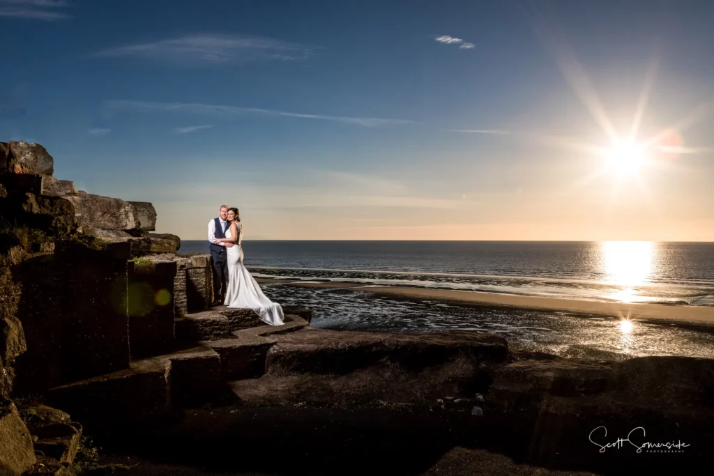 A bride and groom stand together on rocky ruins by the seaside at sunset, with the sun low over the horizon and the sea in the background. Copyright Somerside Photography Ltd // www.somersidephotography.com