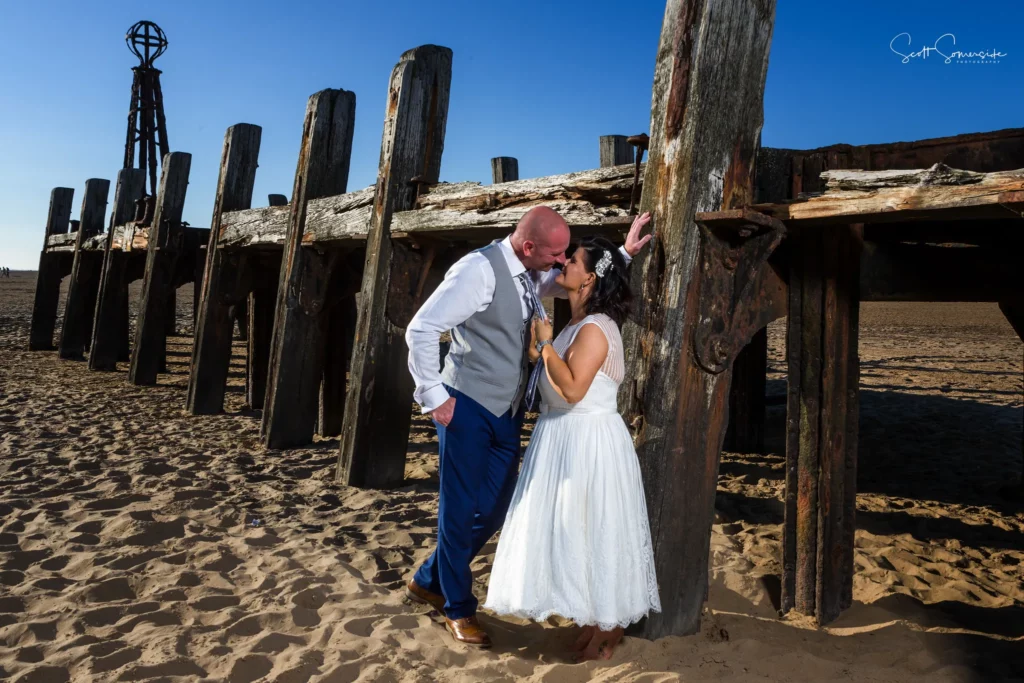 A bride and groom stand close together on a sandy beach near weathered wooden posts, sharing a joyful moment under a clear blue sky. Copyright Somerside Photography Ltd // www.somersidephotography.com