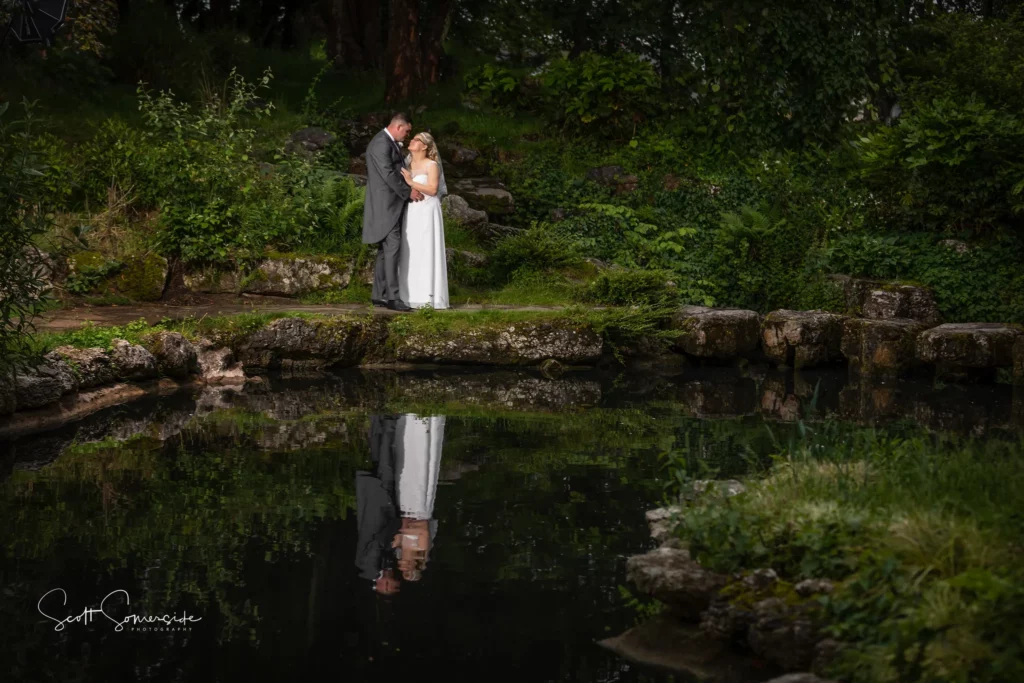A bride and groom stand close together on a stone path beside a pond, with their reflection visible in the water, surrounded by green foliage. Copyright Somerside Photography Ltd // www.somersidephotography.com