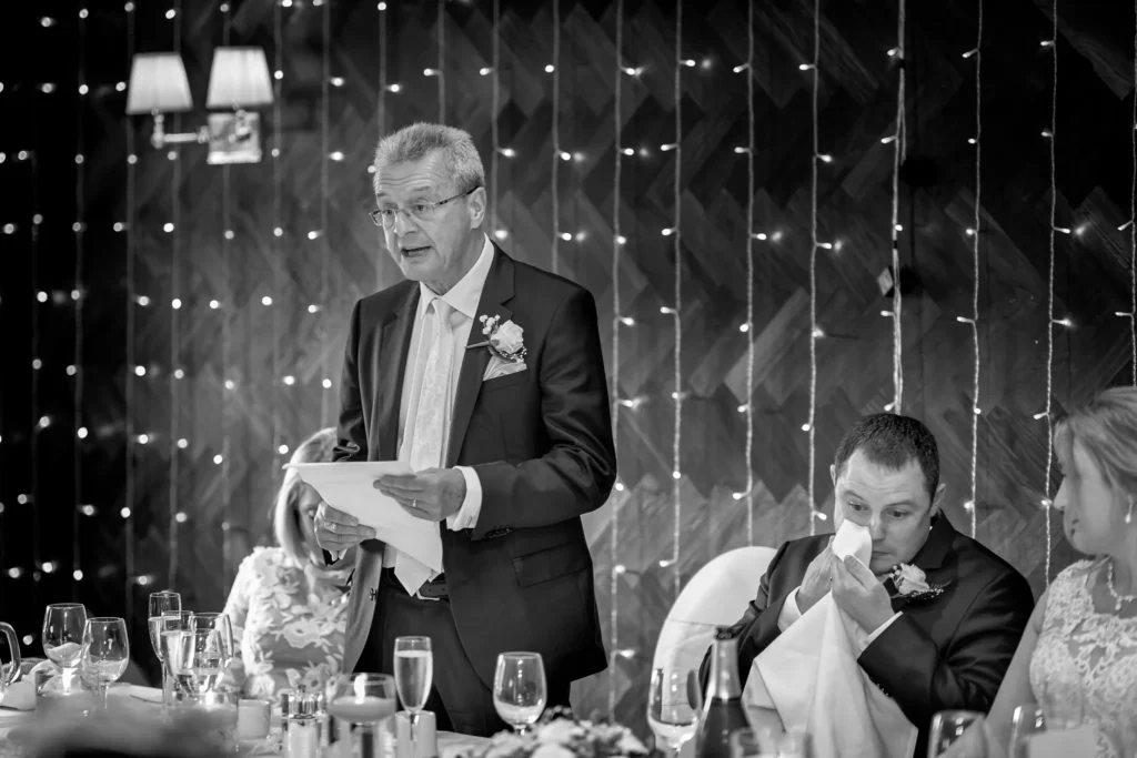 A man in a suit stands and gives a speech at a formal event whilst another man sits beside him, wiping his face with a serviette. Copyright Somerside Photography Ltd // www.somersidephotography.com
