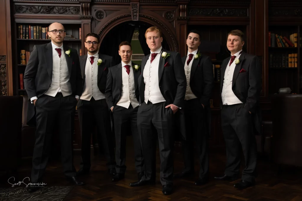 Six men in formal suits with waistcoats and red ties stand together in a library with dark wood bookshelves and ornate decor. Copyright Somerside Photography Ltd // www.somersidephotography.com