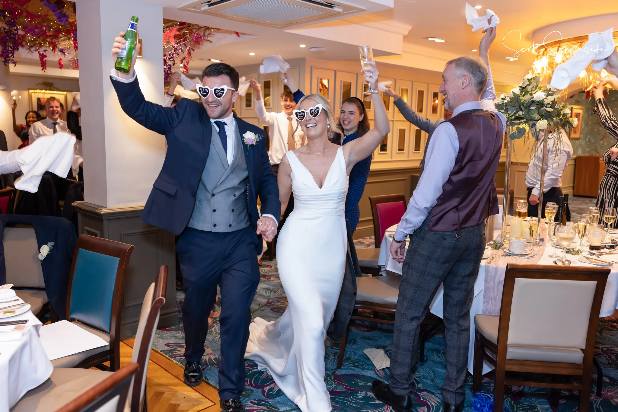 A bride and groom wearing heart-shaped sunglasses enter a wedding reception, smiling and raising drinks, as guests cheer around them. Copyright Somerside Photography Ltd // www.somersidephotography.com
