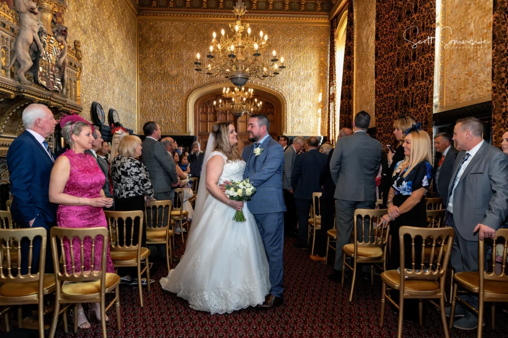 A bride and groom stand facing each other in a grand room, holding hands and smiling, surrounded by seated and standing wedding guests. Copyright Somerside Photography Ltd // www.somersidephotography.com