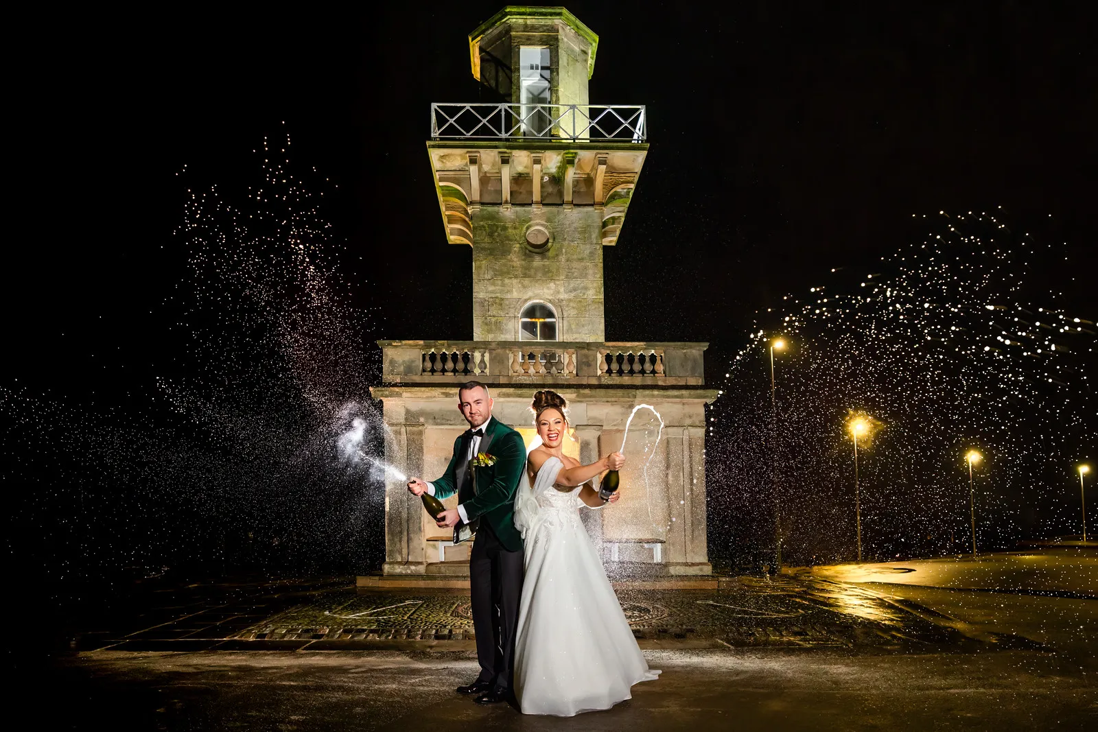 A bride and groom stand in front of a lit stone tower at night, spraying champagne into the air and smiling. Copyright Somerside Photography Ltd // www.somersidephotography.com