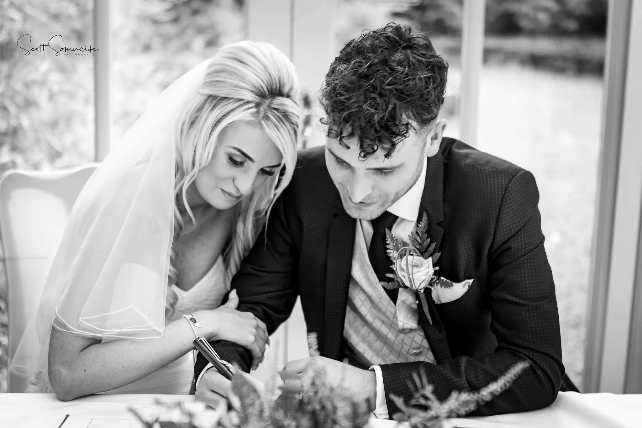 A bride leans on the groom’s shoulder as he signs a document at a table, both dressed in wedding attire. Copyright Somerside Photography Ltd // www.somersidephotography.com