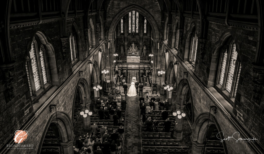 A bride and groom stand at the altar in a grand, stone church, surrounded by guests seated in pews, viewed from a high vantage point. Copyright Somerside Photography Ltd // www.somersidephotography.com