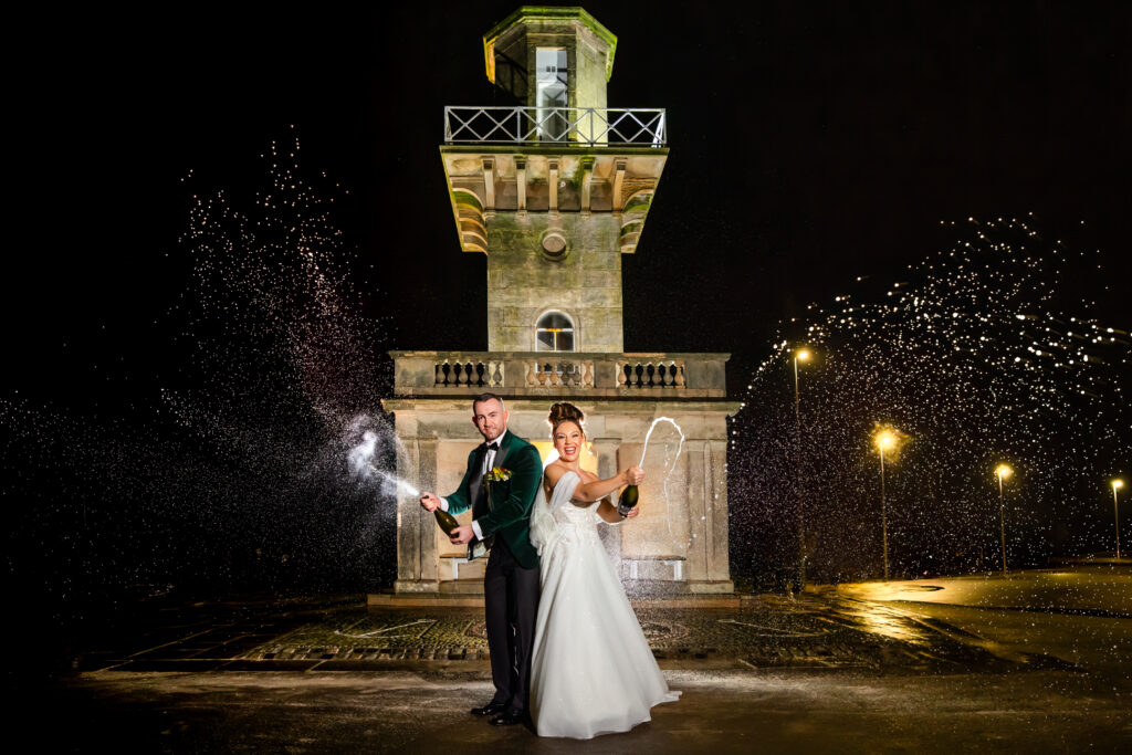 A bride and groom in formal attire stand in front of a tower at night, both spraying champagne, with droplets captured mid-air. Copyright Somerside Photography Ltd // www.somersidephotography.com