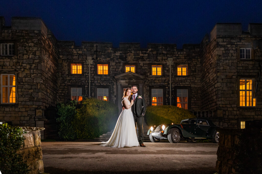 A bride and groom stand together at night in front of a large stone building with lit windows and a vintage car parked nearby. Copyright Somerside Photography Ltd // www.somersidephotography.com