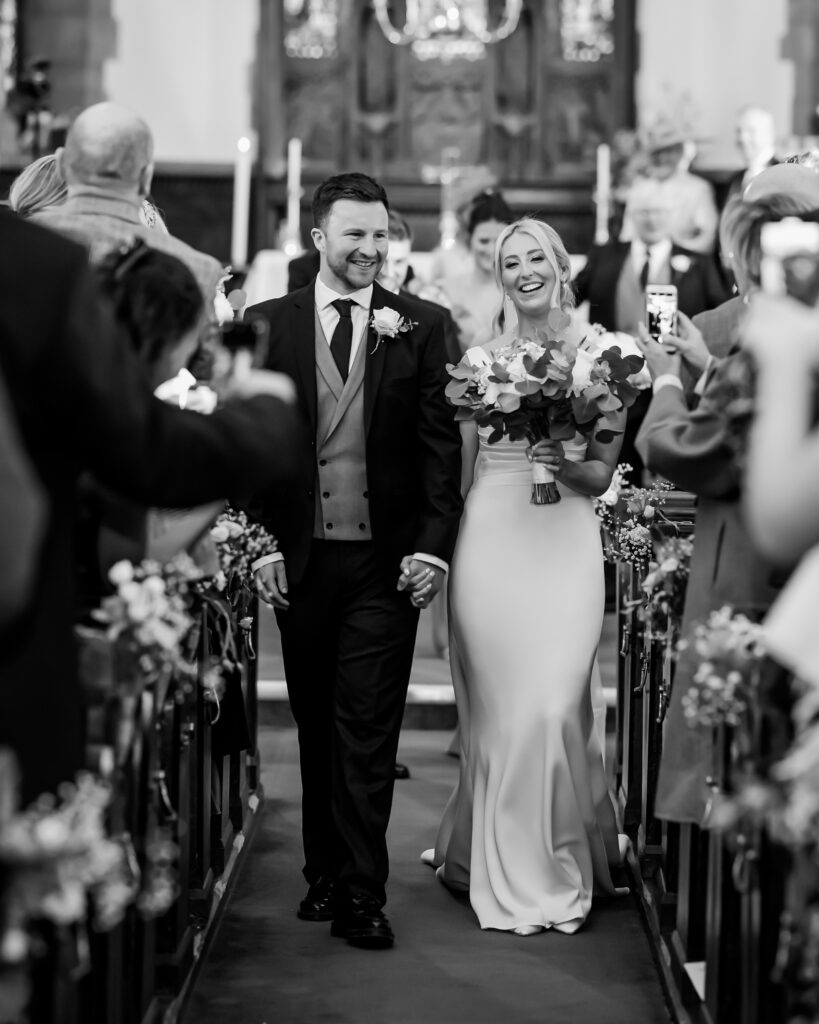 A bride and groom walk down the aisle, smiling and holding hands, as guests take photos and celebrate in a church setting. Copyright Somerside Photography Ltd // www.somersidephotography.com