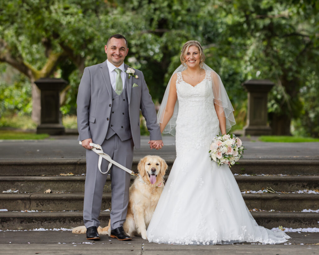 A bride and groom in wedding attire stand outdoors holding hands, with a golden retriever sitting beside them. The bride holds a bouquet and all three face the camera and smile. Copyright Somerside Photography Ltd // www.somersidephotography.com