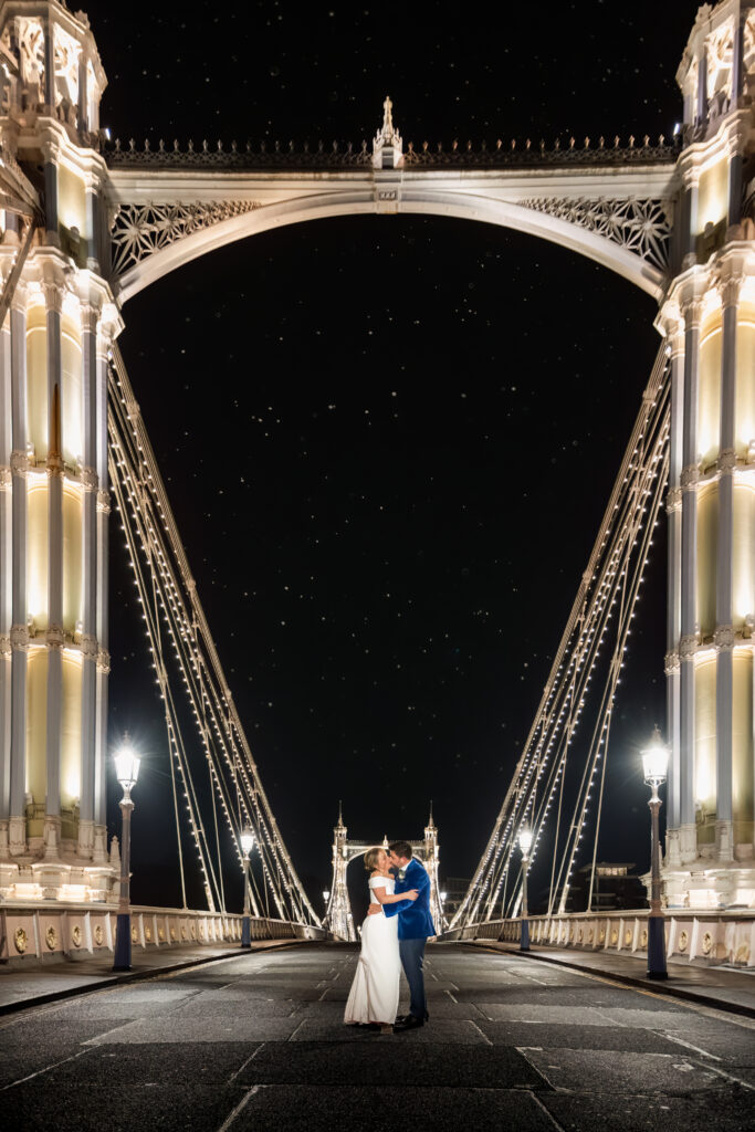 A couple in formal attire stands embracing on a brightly lit suspension bridge at night, with a star-filled sky in the background. Copyright Somerside Photography Ltd // www.somersidephotography.com