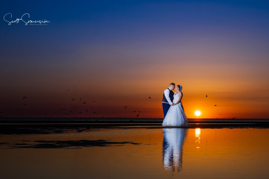 A bride and groom stand together on a beach at sunset, holding hands and reflected in the water, with birds flying in the sky. Copyright Somerside Photography Ltd // www.somersidephotography.com