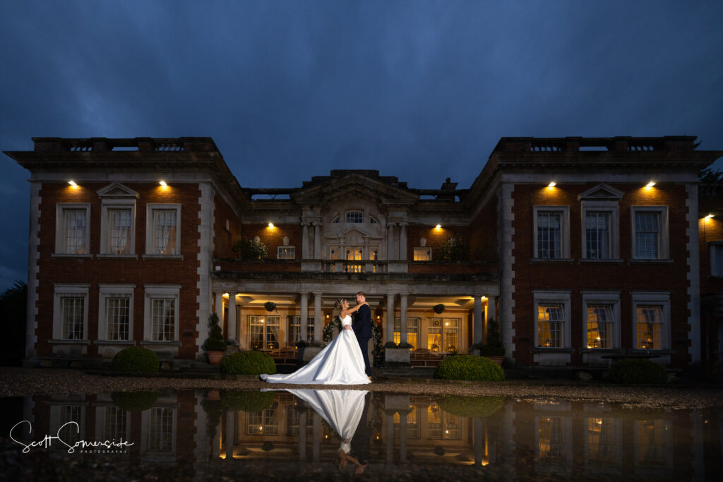 A bride and groom stand together outside an illuminated manor at night, with their reflection visible in a pool of water. Copyright Somerside Photography Ltd // www.somersidephotography.com