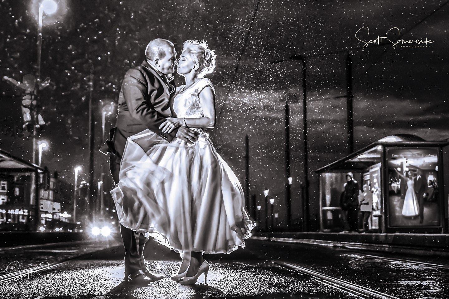 A couple in wedding attire embraces and kisses on a rainy street at night, illuminated by headlights and streetlights, with reflections visible in a glass shelter. Copyright Somerside Photography Ltd // www.somersidephotography.com