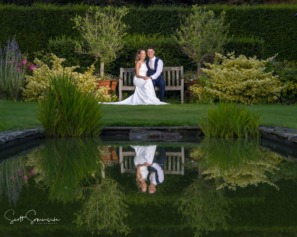 A bride and groom sit on a wooden bench in a garden, with their reflection visible in a pond in the foreground. Copyright Somerside Photography Ltd // www.somersidephotography.com