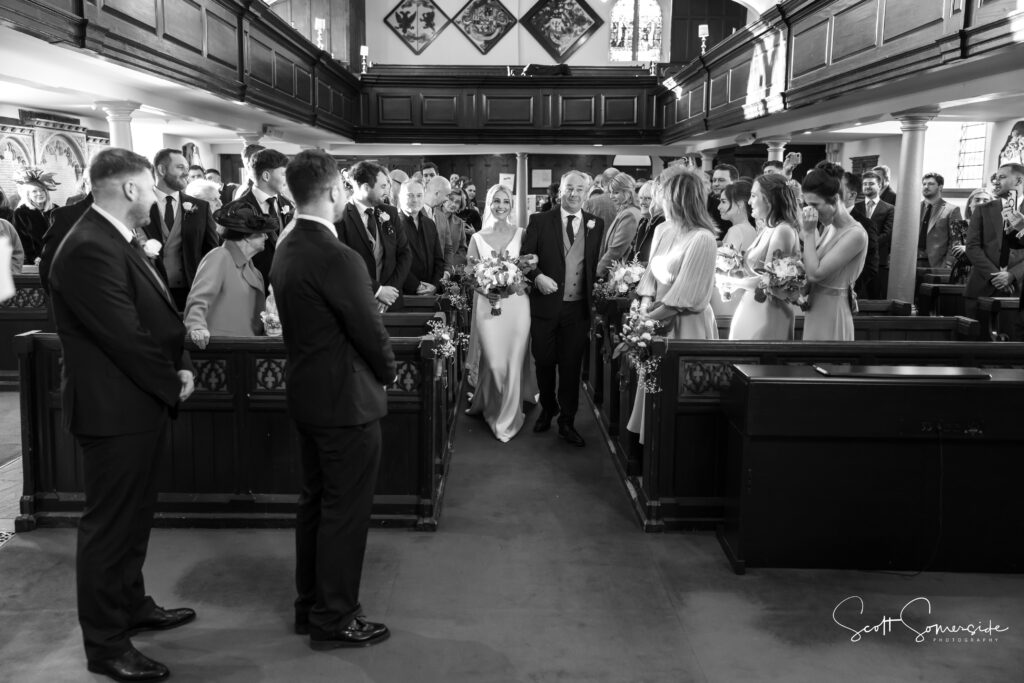 A bride walks down the aisle with an older man in a church, surrounded by standing guests and members of the wedding party, all attentively watching. Copyright Somerside Photography Ltd // www.somersidephotography.com