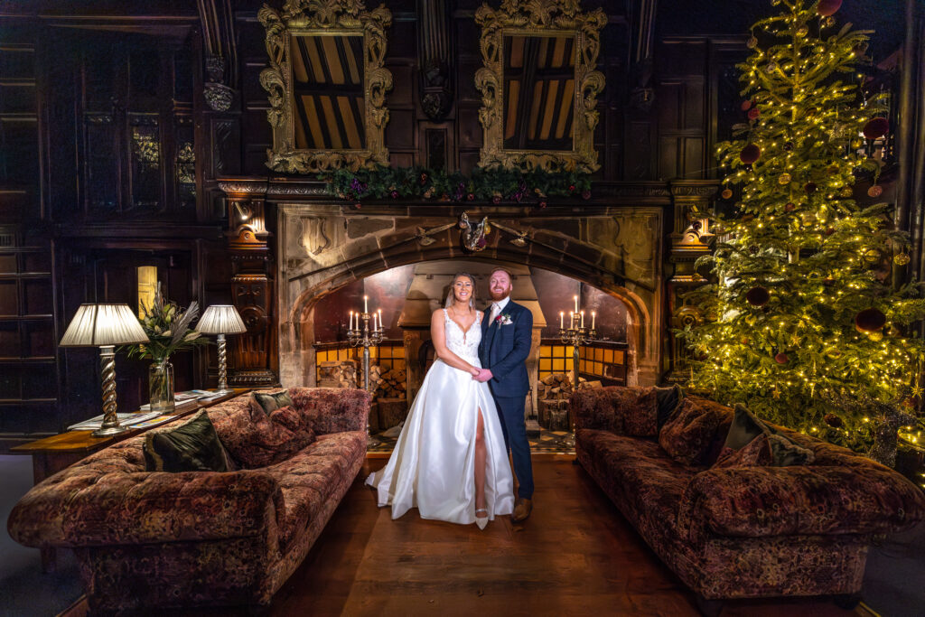A newlywed couple stand together in front of a large fireplace, flanked by sofas and a decorated Christmas tree in an ornately decorated room. Copyright Somerside Photography Ltd // www.somersidephotography.com