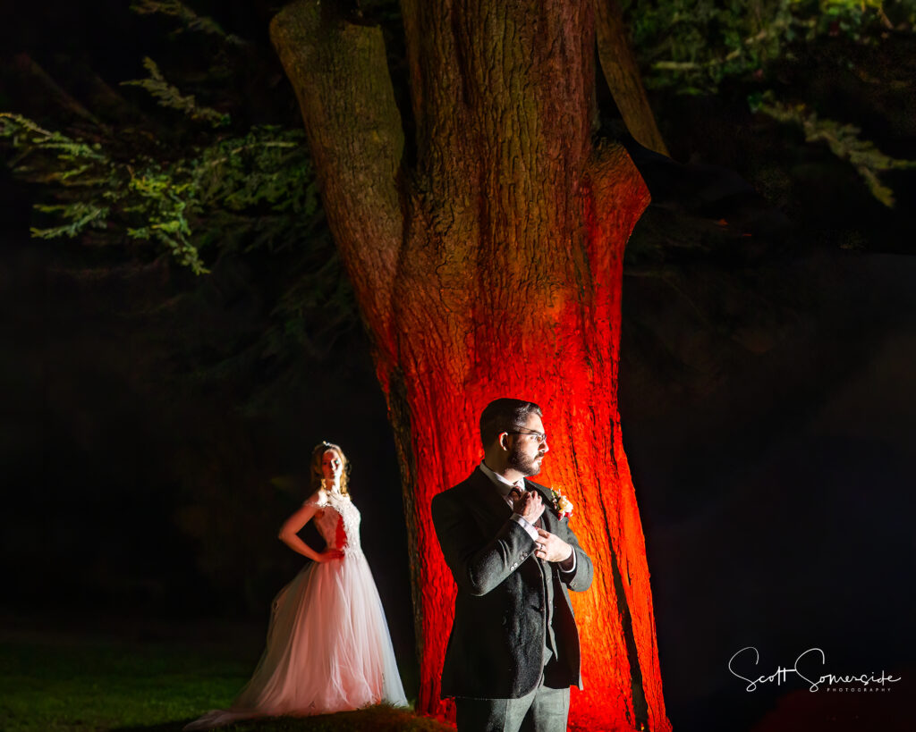 A bride in a white dress stands near a large tree illuminated by red light at night, while a groom in a suit stands in the foreground adjusting his tie. Copyright Somerside Photography Ltd // www.somersidephotography.com