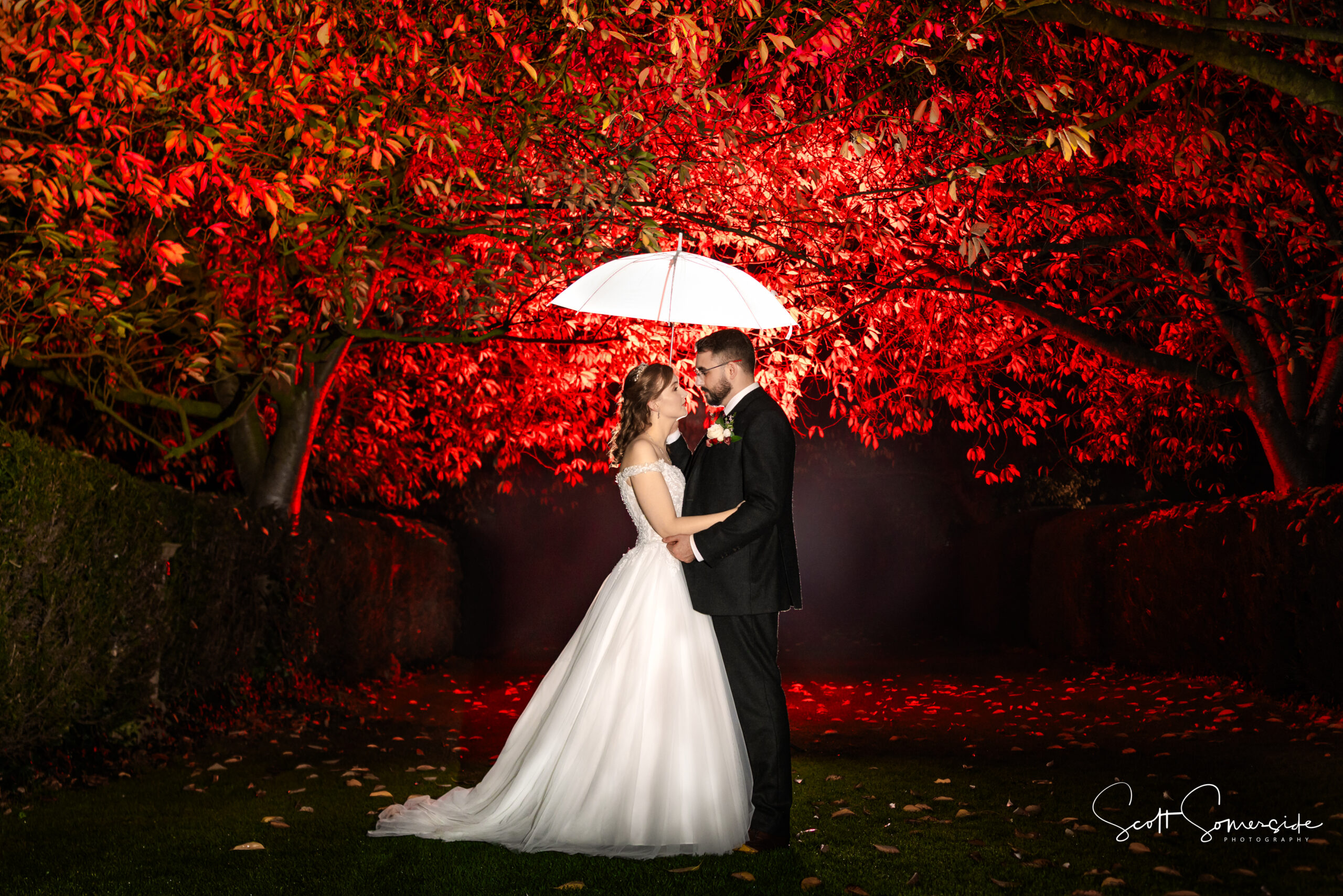 A bride and groom stand under a white umbrella, facing each other at night, surrounded by trees illuminated with red lighting. Copyright Somerside Photography Ltd // www.somersidephotography.com