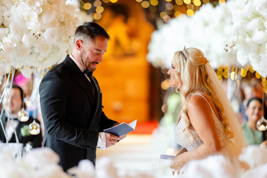 A man and woman stand facing each other at a decorated indoor wedding ceremony, holding papers and smiling, surrounded by white flowers and guests. Copyright Somerside Photography Ltd // www.somersidephotography.com