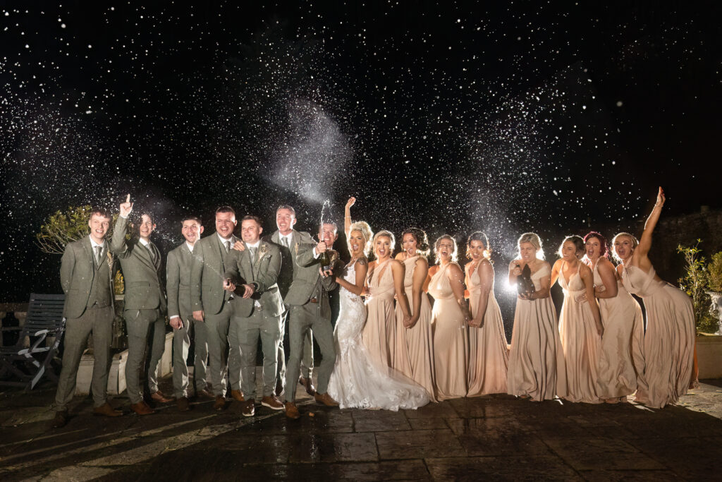A wedding party poses outdoors at night, dressed in formal attire, with confetti or champagne spraying in the air behind them. Copyright Somerside Photography Ltd // www.somersidephotography.com