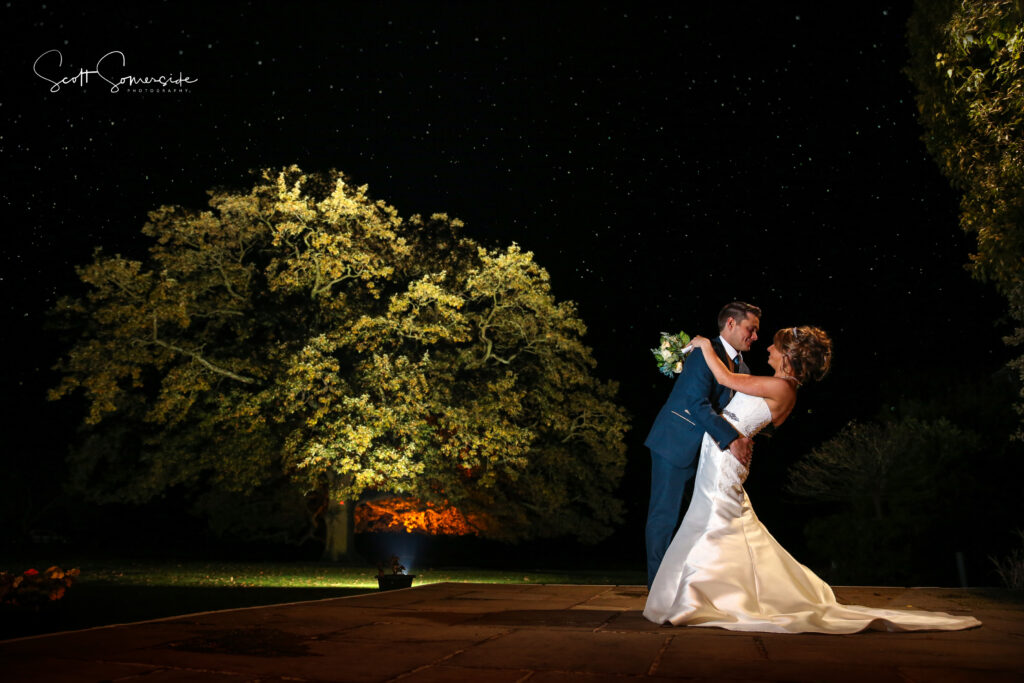 A bride and groom pose in a dip on an outdoor stone patio at night, with a large illuminated tree and starry sky in the background. Copyright Somerside Photography Ltd // www.somersidephotography.com