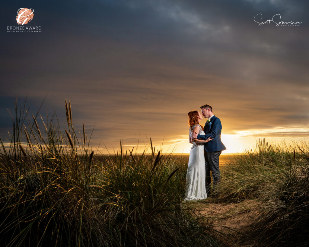 A bride and groom stand close together in tall grass at sunset, under a dramatic cloudy sky. Copyright Somerside Photography Ltd // www.somersidephotography.com