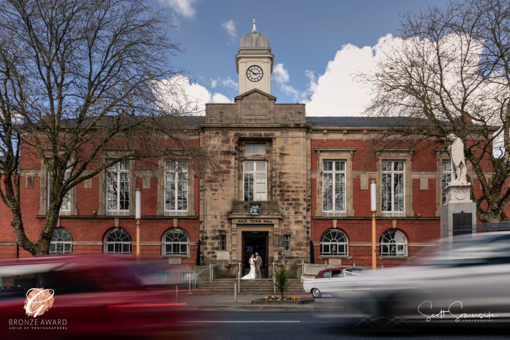 A red-brick town hall with a clock tower and stone entrance is seen with blurred cars passing in the foreground and two people standing at the entrance. Copyright Somerside Photography Ltd // www.somersidephotography.com