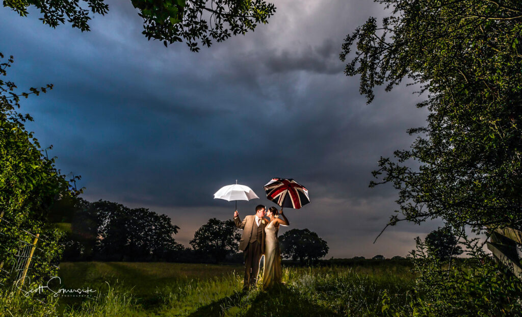 A couple stand together in a grassy field at dusk, holding a white umbrella and a Union Jack umbrella, surrounded by trees under a dramatic, cloudy sky. Copyright Somerside Photography Ltd // www.somersidephotography.com