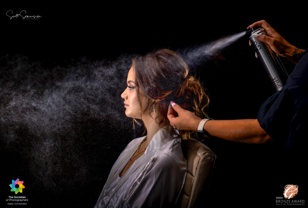 A woman in a white dressing gown sits in profile while someone sprays her styled hair with hairspray against a black background. Copyright Somerside Photography Ltd // www.somersidephotography.com