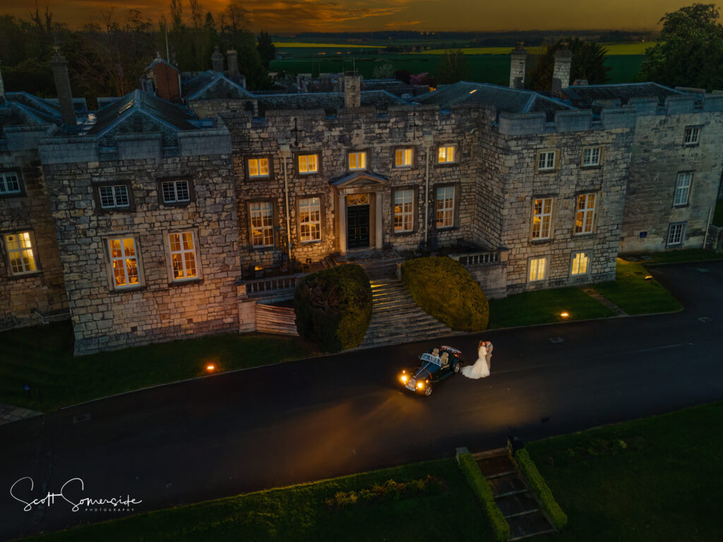 A bride and groom stand beside a vintage car in front of an illuminated stone mansion at sundown. Copyright Somerside Photography Ltd // www.somersidephotography.com