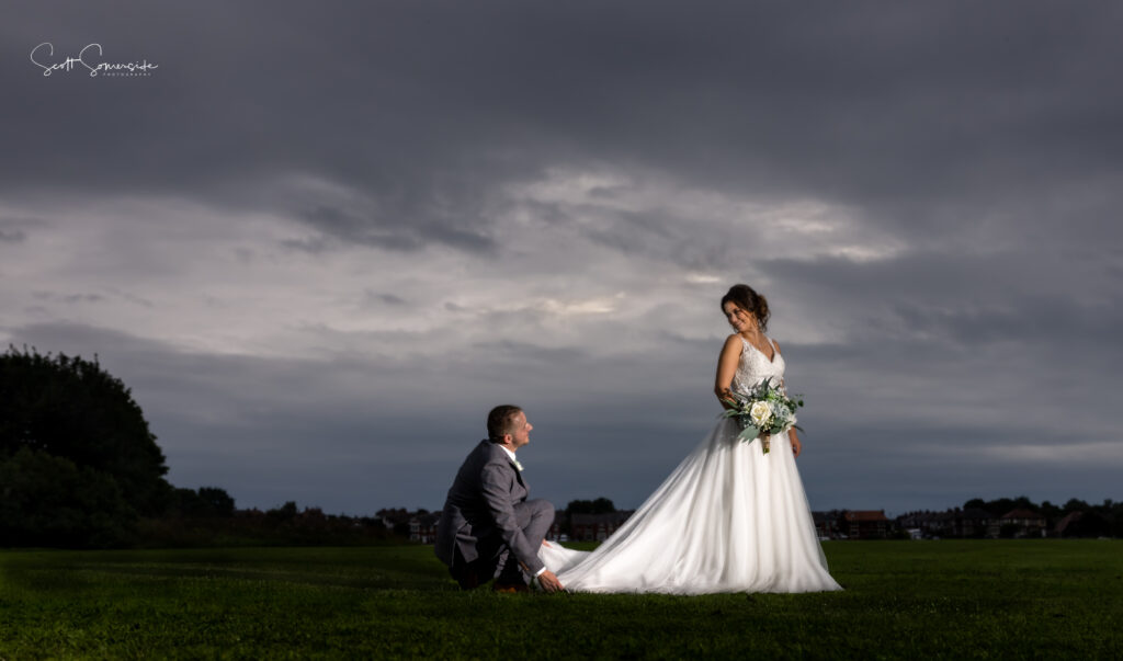 A groom kneels on grass, holding the bride’s dress train, while the bride stands holding a bouquet; both are outdoors under a cloudy sky. Copyright Somerside Photography Ltd // www.somersidephotography.com