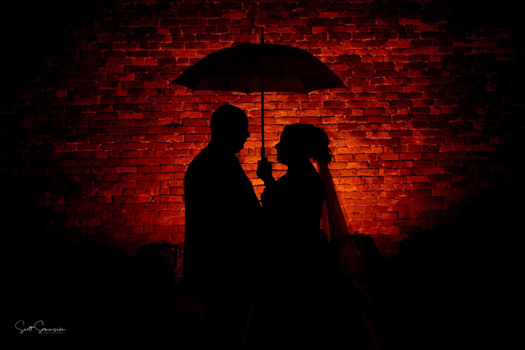 Silhouette of a couple under an umbrella in front of a red-lit brick wall. Copyright Somerside Photography Ltd // www.somersidephotography.com