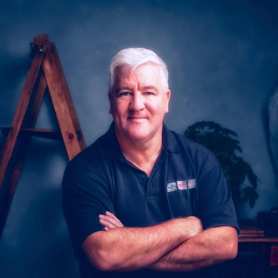 A middle-aged man with short white hair, wearing a dark polo shirt, stands with arms folded in front of a ladder and a plant, against a blue background. Copyright Somerside Photography Ltd // www.somersidephotography.com